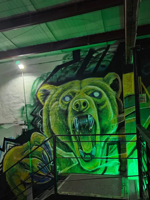 Oso verde y amarillo de ojos celestes brillantes y fauces abiertas, pintado en la pared de un gimnasio, con la baranda de la escalera metálica delante y luz verde sobre el techo.