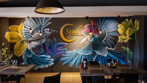 Mural de dos pájaros azules con alas abiertas y flores amarillas y violetas a ambos lados del cartel dorado del restaurante, sobre una pared oscura, con mesas puestas delante y luz cálida.