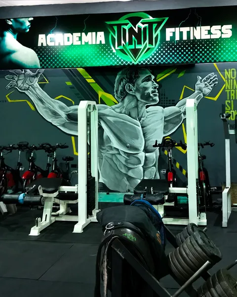 Mural en blanco, negro y verde menta de un fisicoculturista con los brazos abiertos y la mirada en alto, bajo un cartel luminoso verde de gimnasio, con máquinas de musculación delante y una frase amarilla a la derecha.