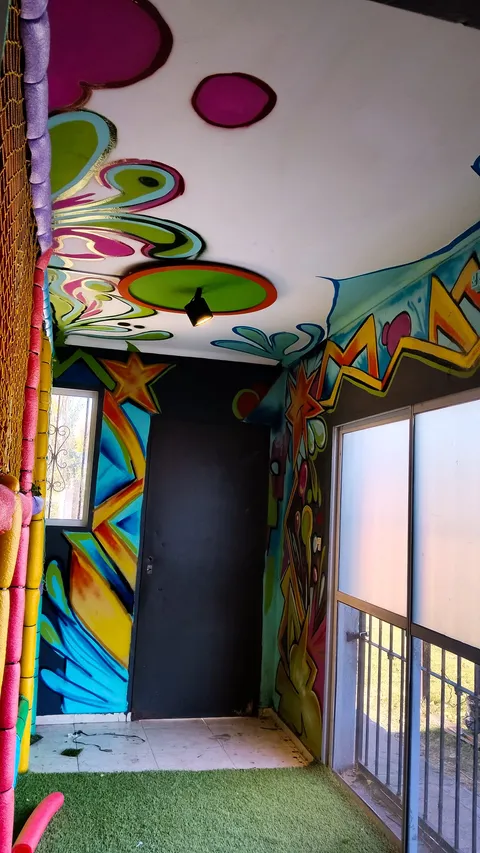 Sala de juegos infantil con paredes y techo pintados con flores magenta, estrellas naranjas y destellos amarillos y azules, sobre piso de césped sintético y una puerta oscura al fondo.