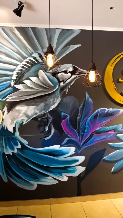 Detalle del pájaro azul de alas abiertas pintado con plumas celestes, grises y blancas, junto a hojas violetas y rosadas, bajo dos lámparas colgantes de filamento.