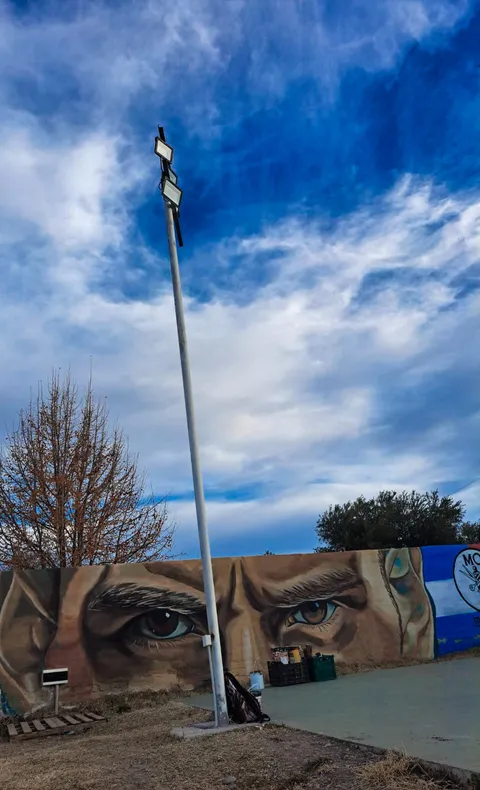 Vista abierta de la cancha con el paredón pintado a lo largo del fondo, un poste de iluminación alto y un cielo azul intenso con nubes.