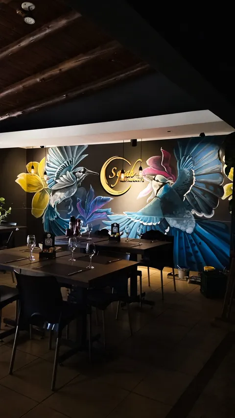 Salón del restaurante con mesas y copas puestas, bajo un techo de vigas de madera, y el mural de pájaros azules iluminado al fondo.