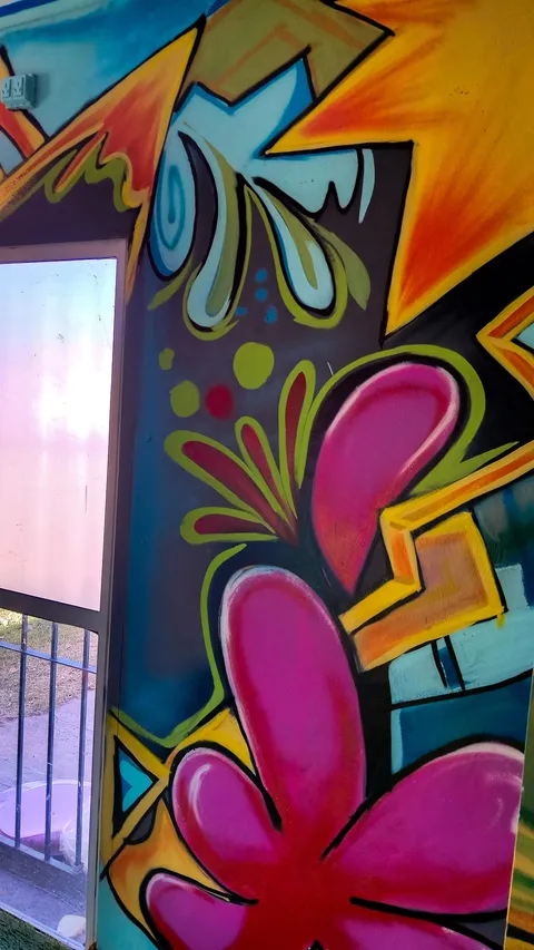 Flor magenta gigante de pétalos rojos con contornos amarillos, pintada en la pared junto a una ventana, entre gotas celestes y formas naranjas.