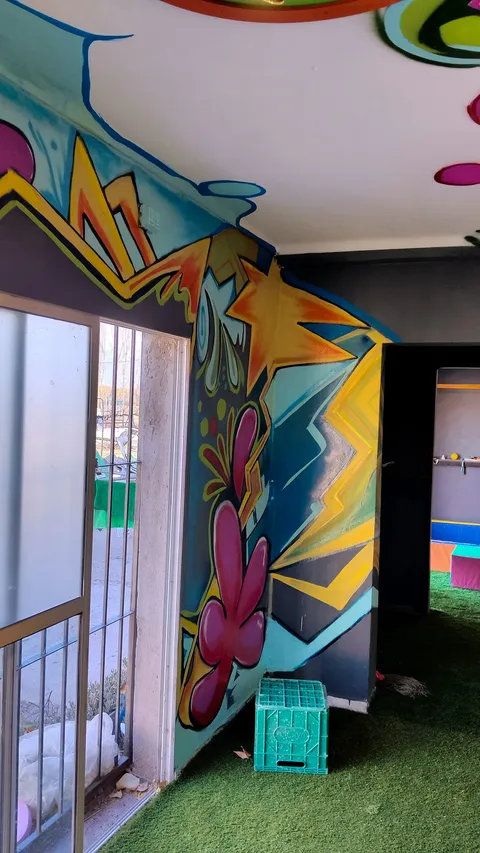 La sala vista desde la entrada: la pared junto al ventanal pintada con flores y zigzags de colores, un banquito verde sobre el césped sintético y el techo con un círculo rojo.