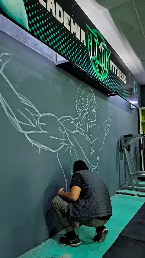 El muralista, agachado y de espaldas, traza con líneas blancas el boceto del mural sobre la pared gris del gimnasio, todavía sin pintar, bajo el cartel verde.