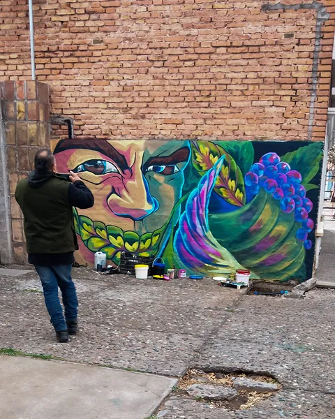Una persona de espaldas pinta el ojo del rostro sobre el muro de ladrillo, con tarros de pintura apoyados en la vereda de baldosas.