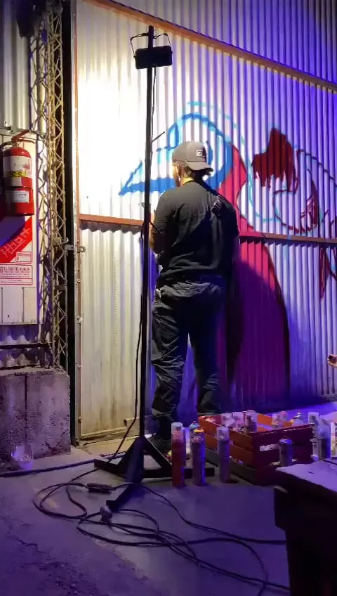 Adri, de espaldas, pinta con aerosol una figura celeste y magenta sobre una pared de chapa, rodeado de latas de aerosol y con luz violeta de noche.