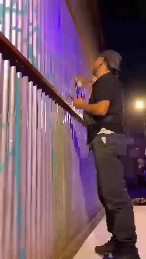 Adri, de perfil y con gorra, pintando con aerosol una pared de chapa iluminada de violeta, de noche.