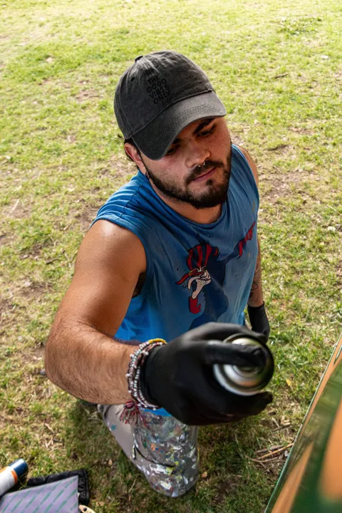 Adri, de barba y gorra gris, arrodillado sobre el pasto con un guante negro, apunta una lata de aerosol hacia la cámara con la mirada atenta; lleva una musculosa azul y pantalón con salpicaduras de pintura.